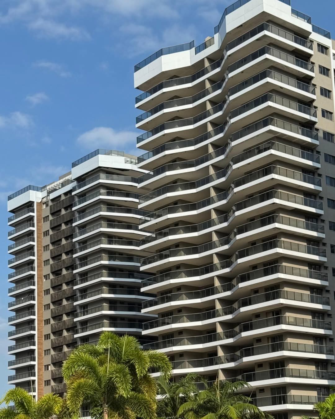 prédios residenciais modernos de alto padrão com varandas envidraçadas em curvas, céu azul e palmeiras na base